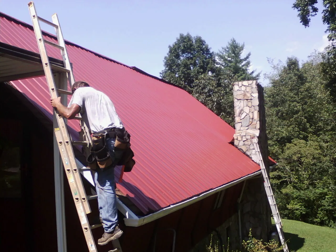 Roofer on a ladder beside a red metal roof and stone chimney — Soffit & Fascia Repair by Roofing Brothers in Elkview